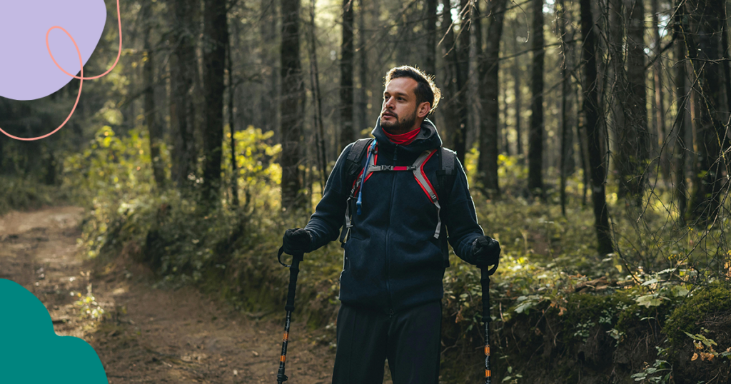 Mies vaeltaa metsässä polulla käyttäen kävelysauvoja. Hänellä on yllään tumma takki ja reppu, ja hän katsoo viistoon vasemmalle. Taustalla näkyy tiheää metsää ja auringonvalo siivilöityy puiden läpi.