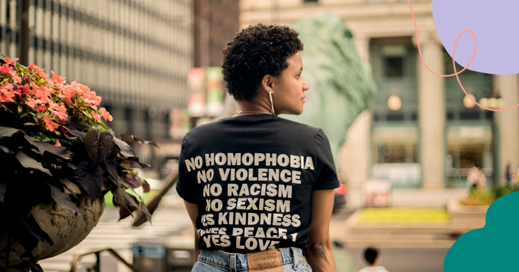 Henkilö seisoo kaupungin kadulla selkä kameraan päin. Hänellä on musta t-paita, jossa lukee valkoisella tekstillä: "No homophobia, No violence, No racism, No sexism, Yes kindness, Yes peace, Yes love". Etualalla vasemmalla on kukkaruukku, jossa on värikkäitä kukkia.