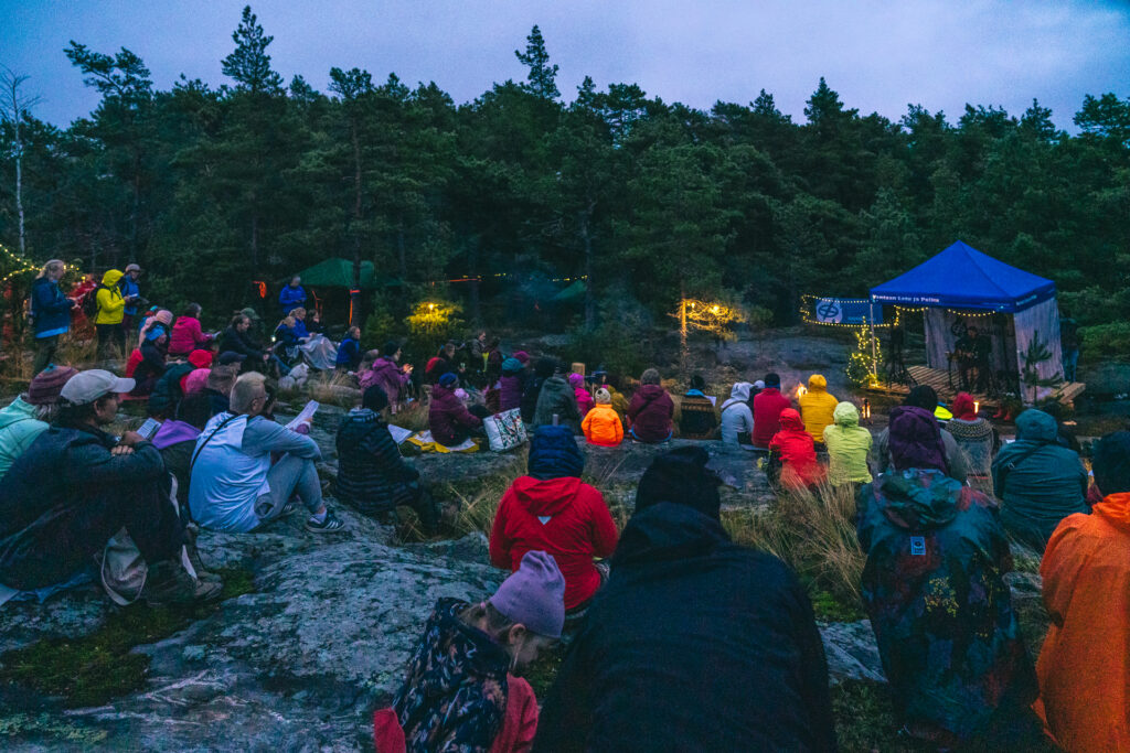 Hämärtyvässä illassa suuri joukko ihmisiä on kokoontunut metsäiseen ulkoilmatapahtumaan. Osallistujat istuvat kallioilla ja sammalikossa värikkäisiin ulkoiluvaatteisiin pukeutuneina ja seuraavat esitystä. Oikealla on sininen esiintymiskatos, jota valaisevat tunnelmalliset valoketjut. Taustalla kohoaa tiheä mäntymetsä.